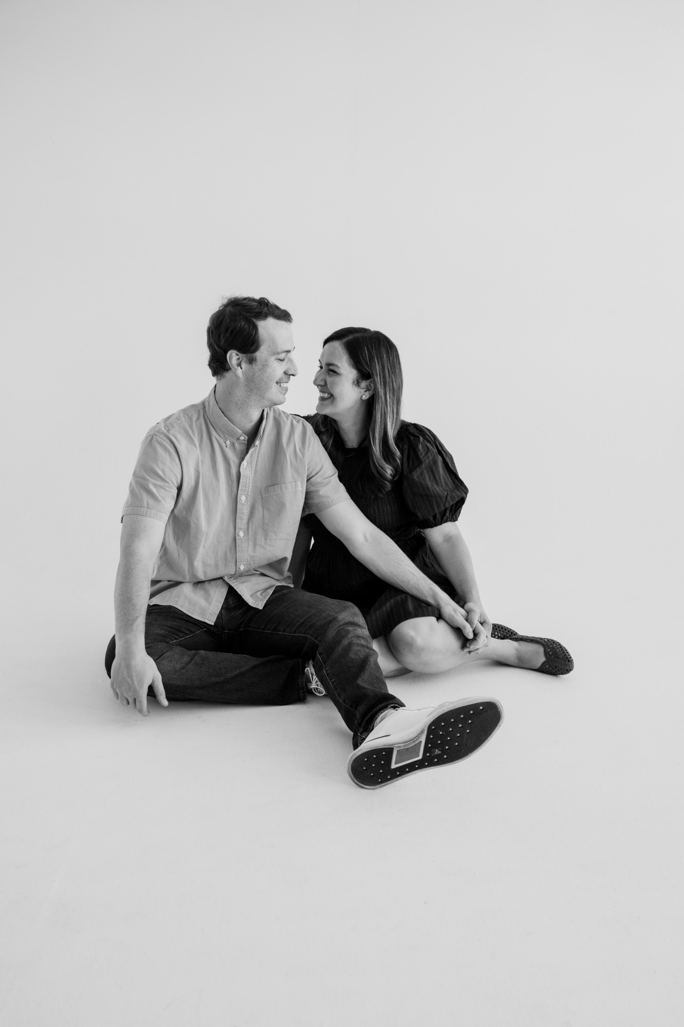 Two people sitting on the floor, facing each other, smiling, with a plain background.