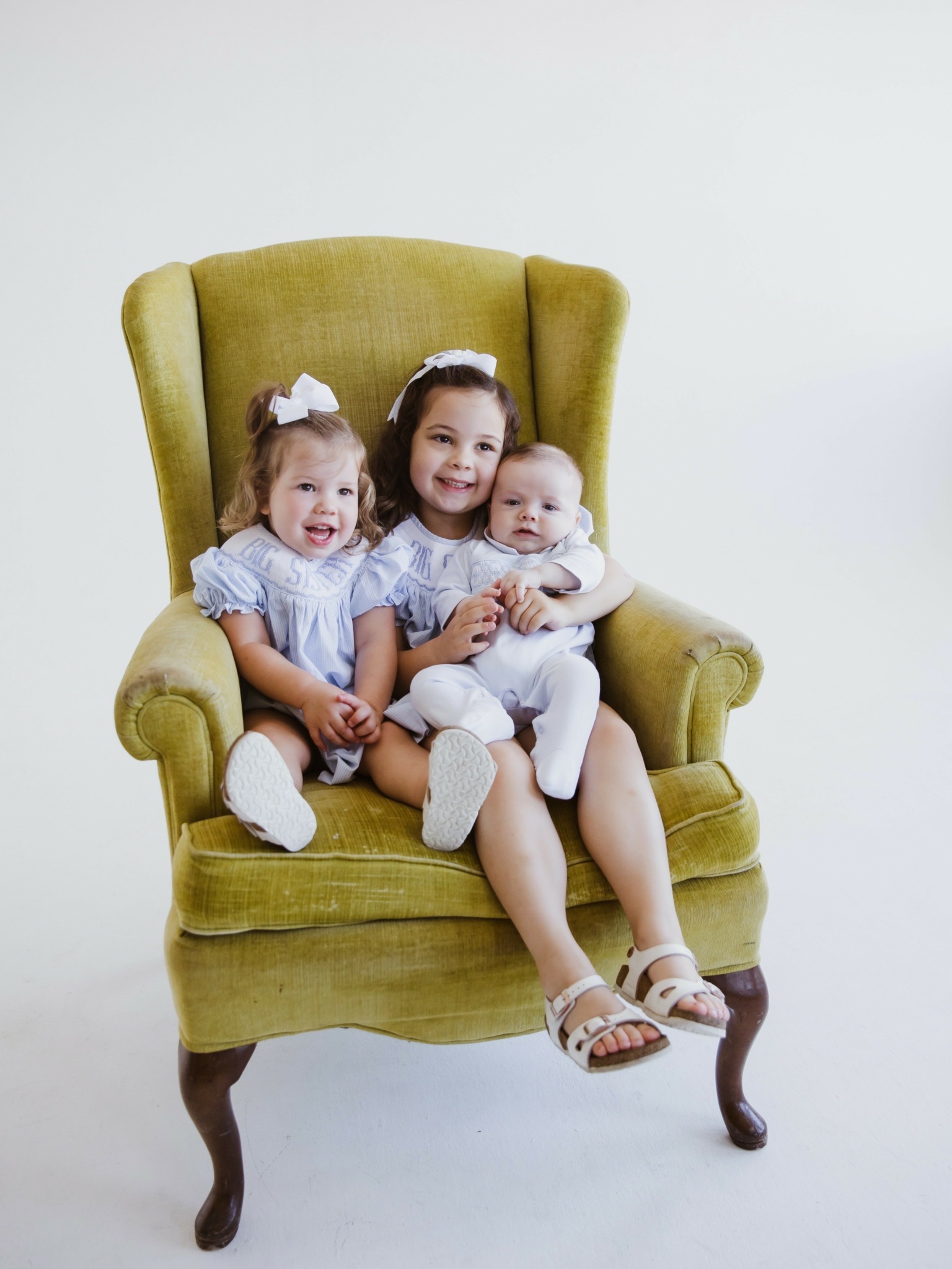 Three children sitting on a green armchair, smiling, with a plain white background.
