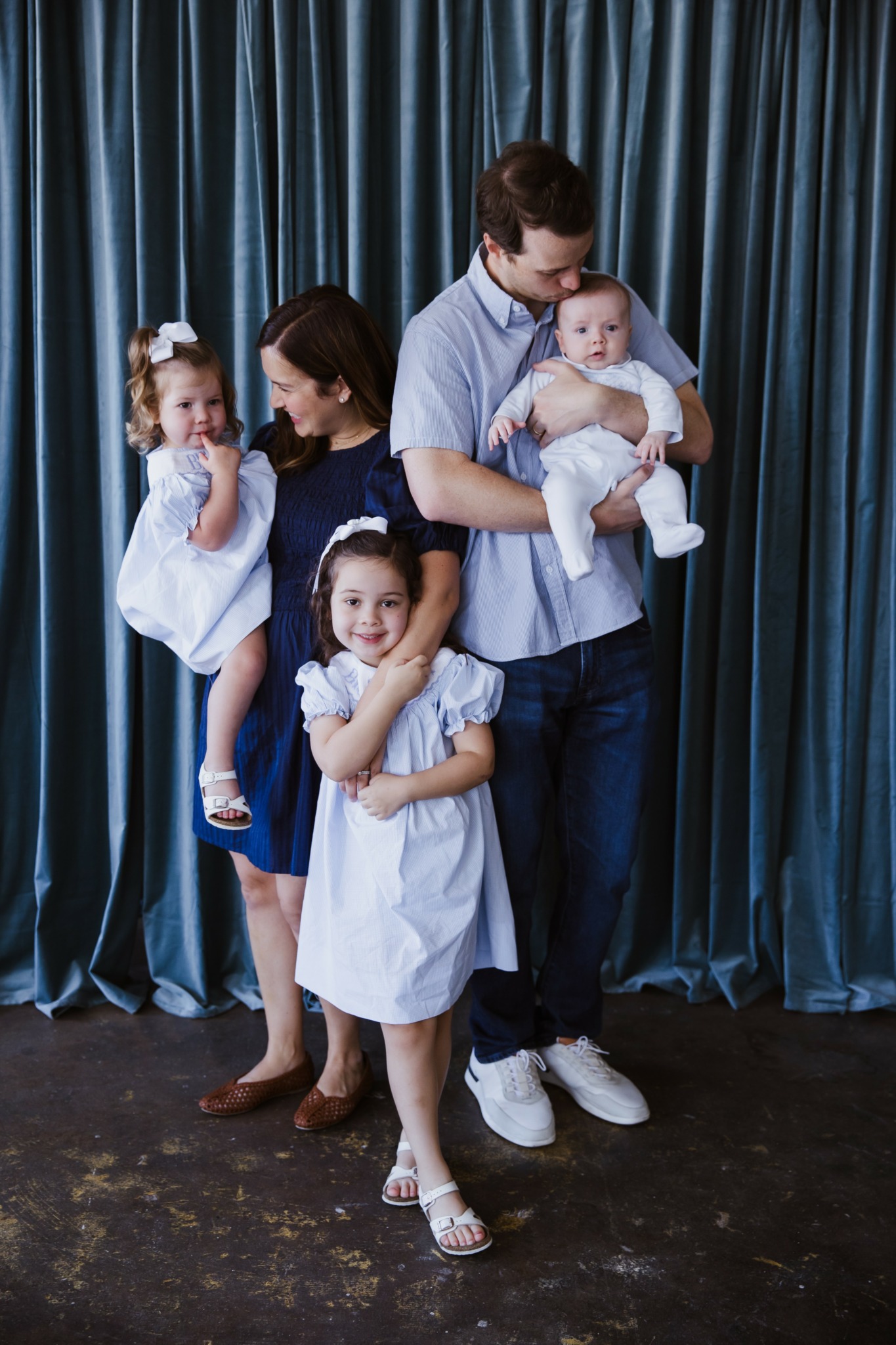 A family of five standing in front of a blue curtain, with children dressed in white and a man holding a baby.