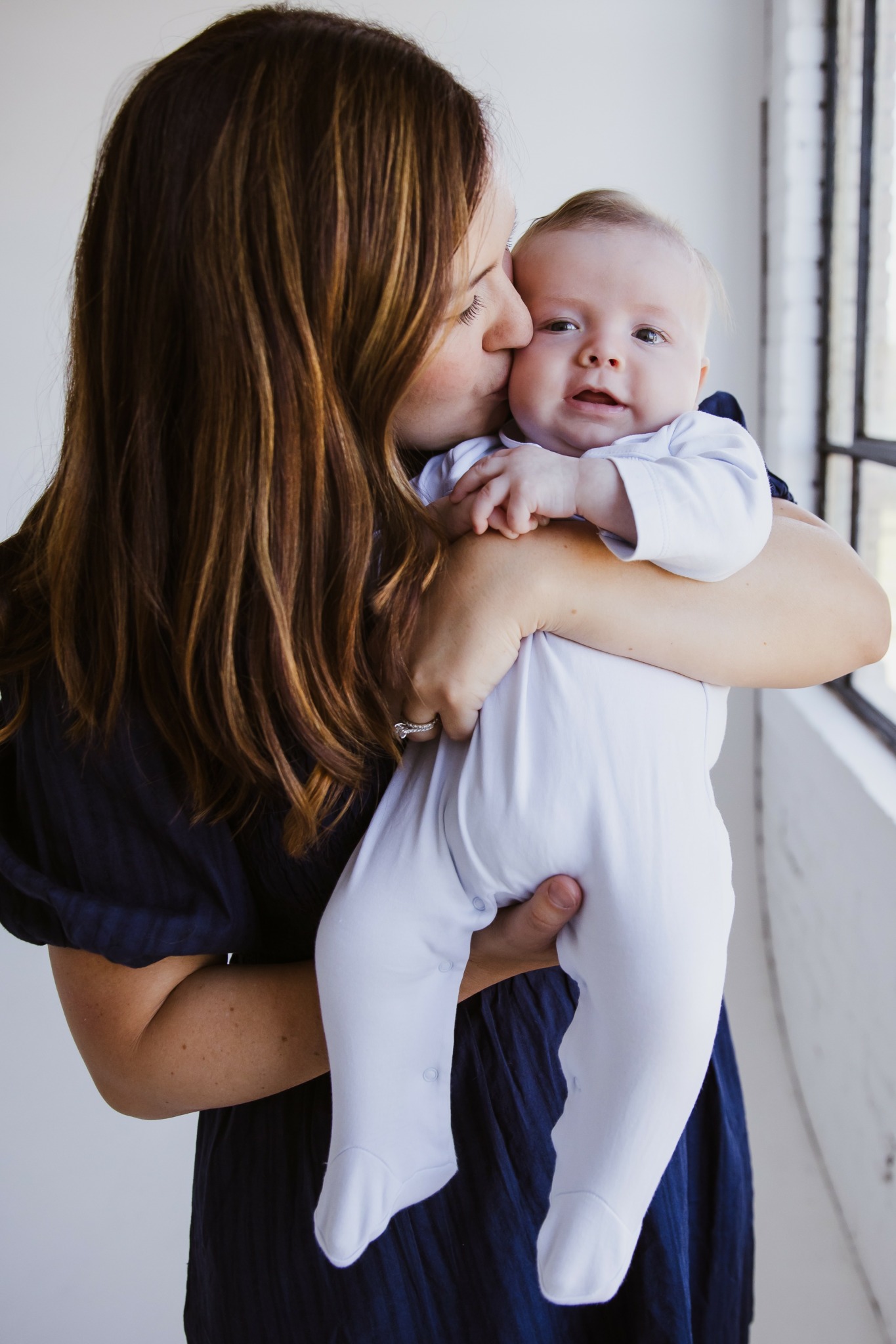 Woman holding a baby near a window, woman kissing baby's cheek, baby looking at camera, light coming through window