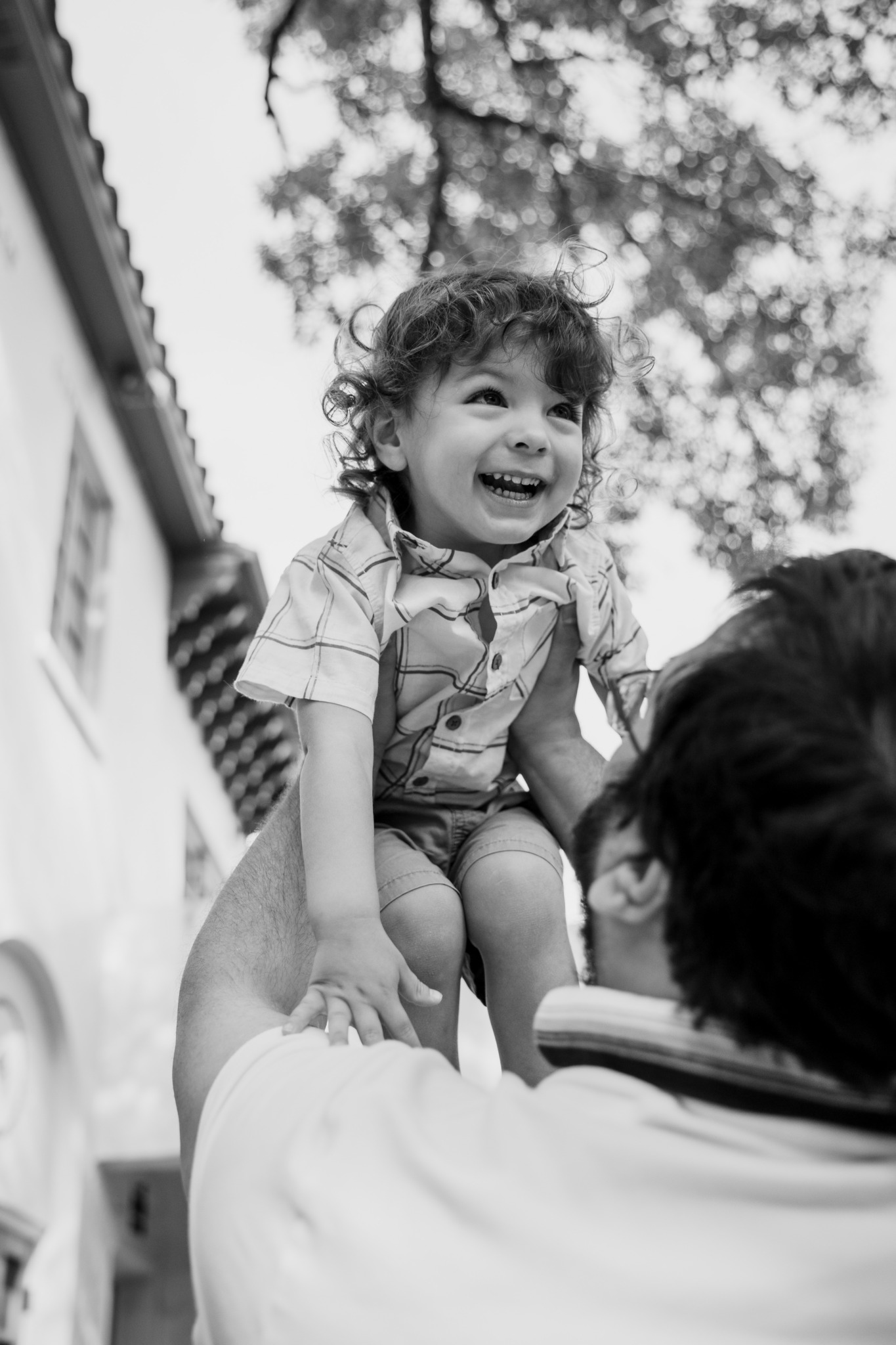 Child with curly hair smiling and being lifted by an adult outdoors, trees and building in background.