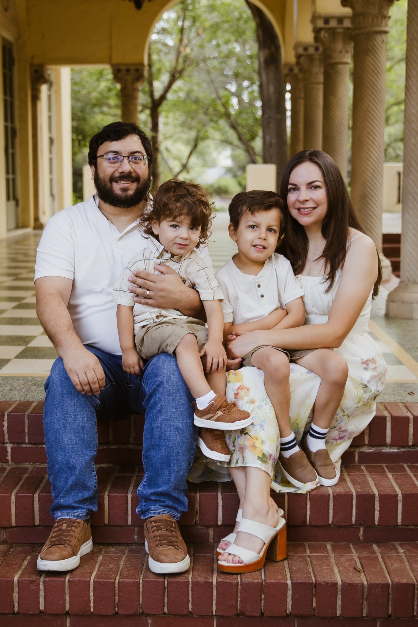 Family of four sitting on steps outdoors, smiling, with trees and columns in background.