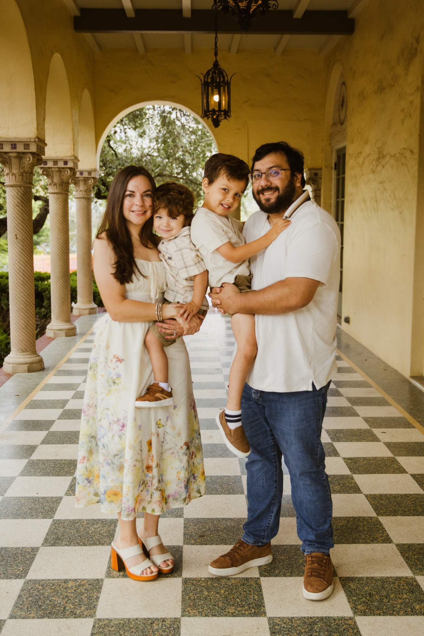 Family of four standing in a covered outdoor corridor with arches and checkered floor, smiling and hugging each other.