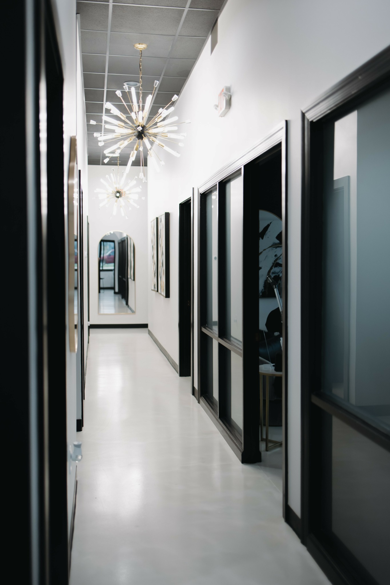 Long hallway with black-framed glass doors on the right, white walls, and a ceiling light fixture, leading to a door at the end.
