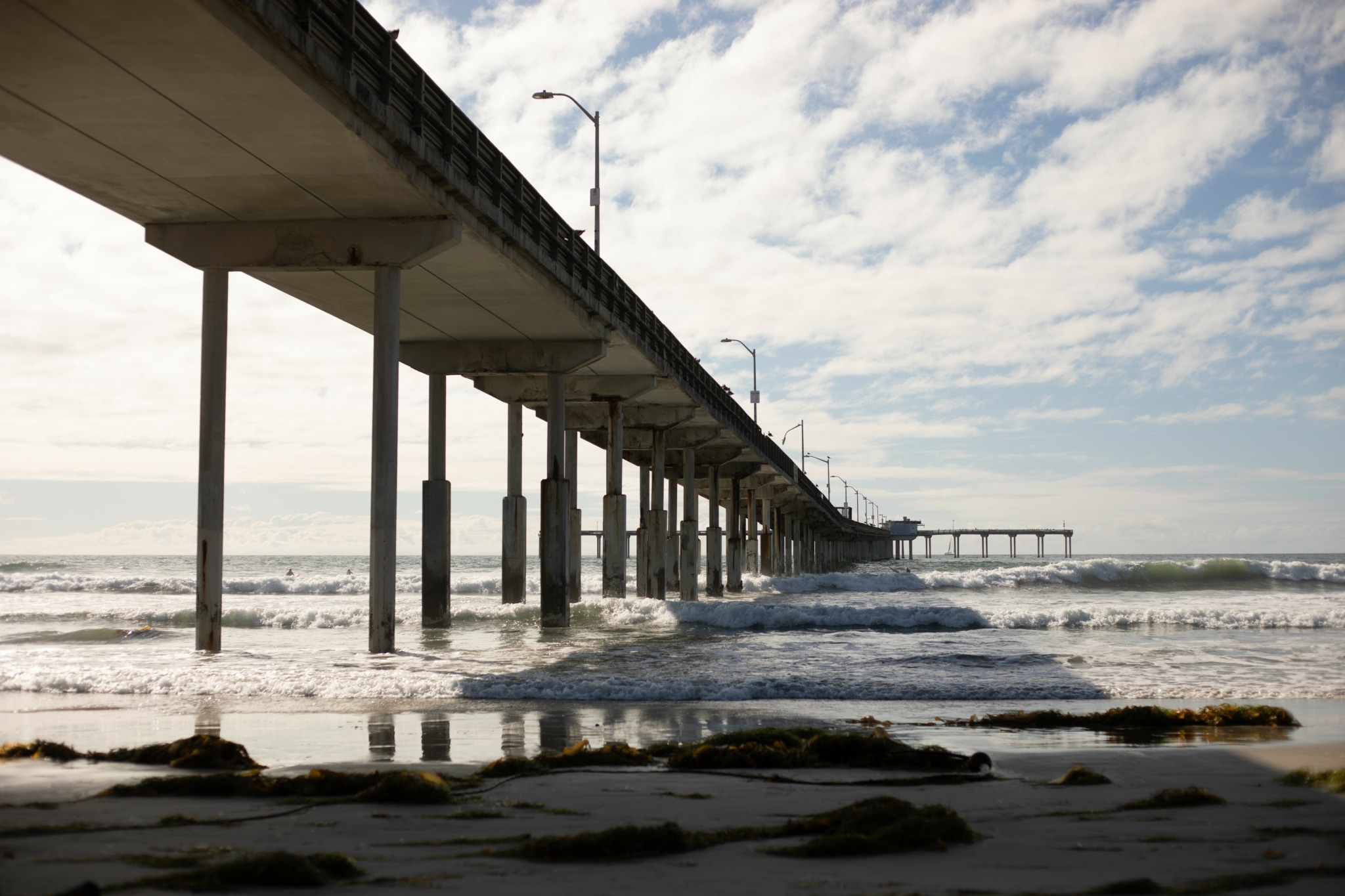 View of a pier extending into the ocean with waves and a partly cloudy sky.