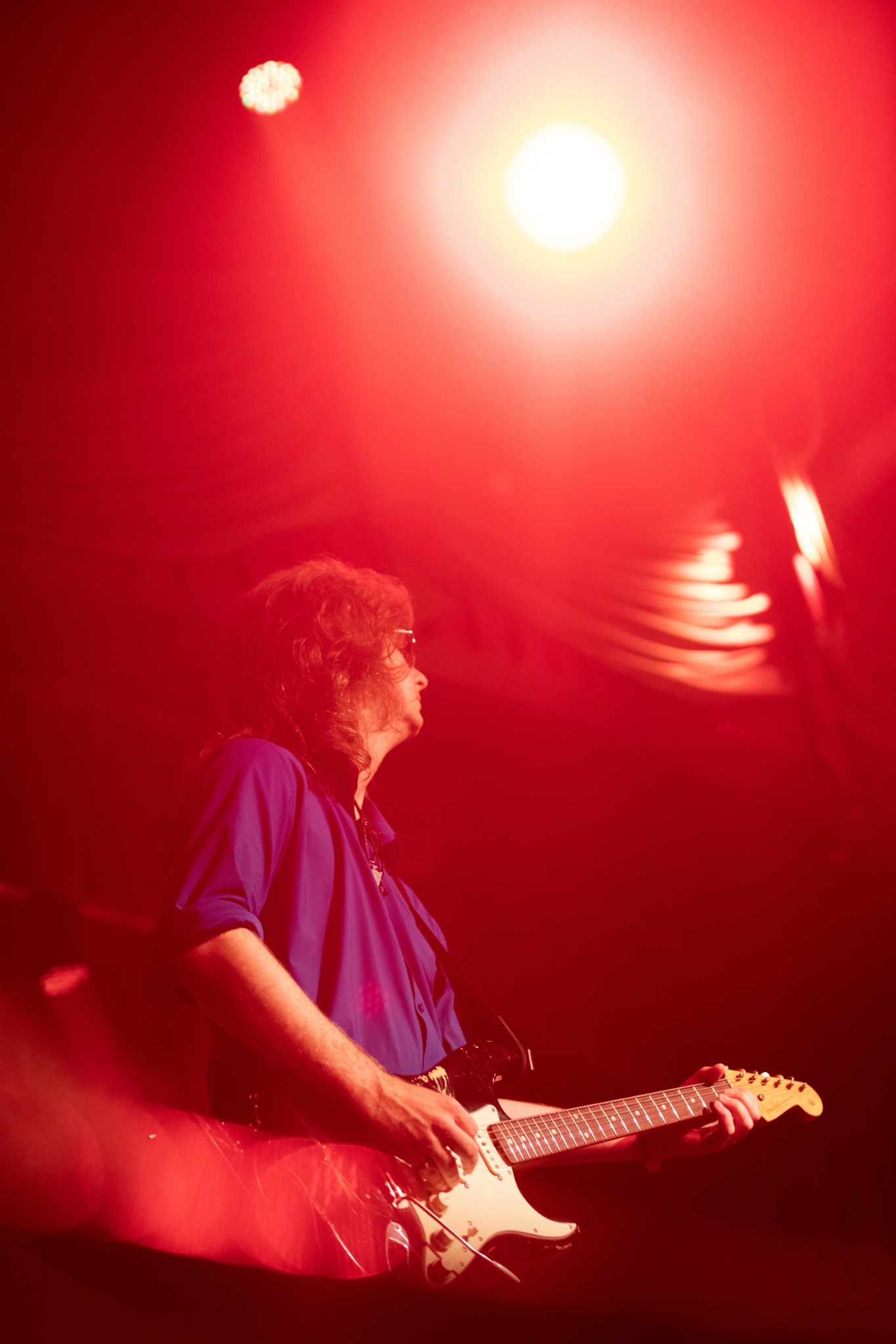 Person playing electric guitar on stage with bright red lighting and reflections, side profile, curly hair, wearing a blue shirt.