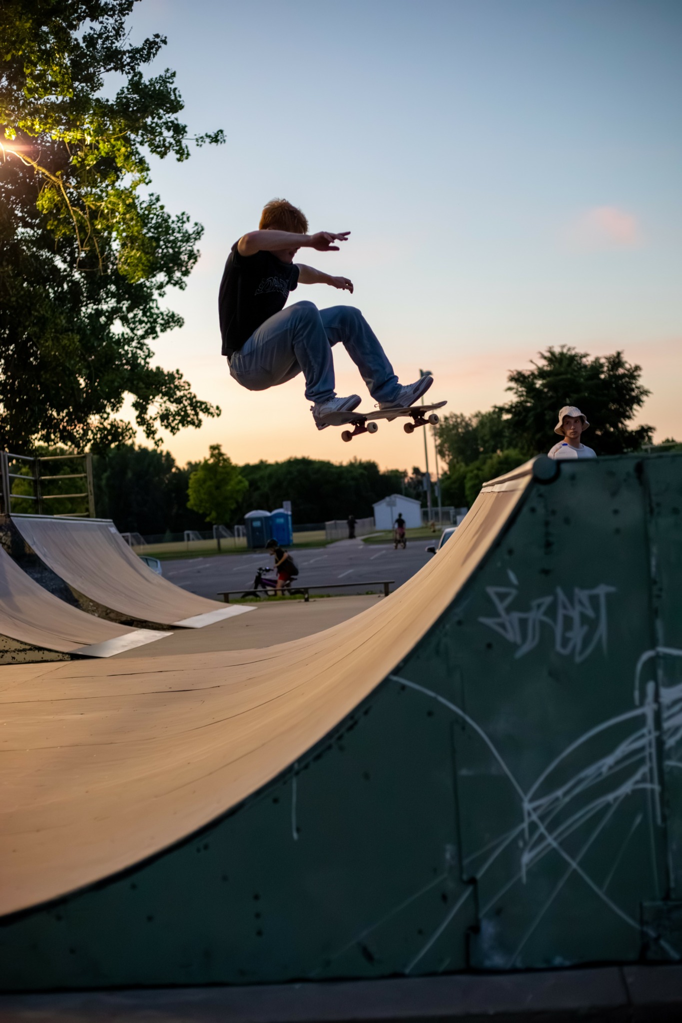 Skateboarder mid-air at skate park with trees and people in background, clear sky, and graffiti on ramp.
