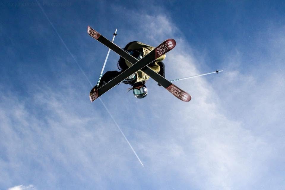 A person skydiving with skis and a helmet, falling through blue sky with clouds, viewed from below.