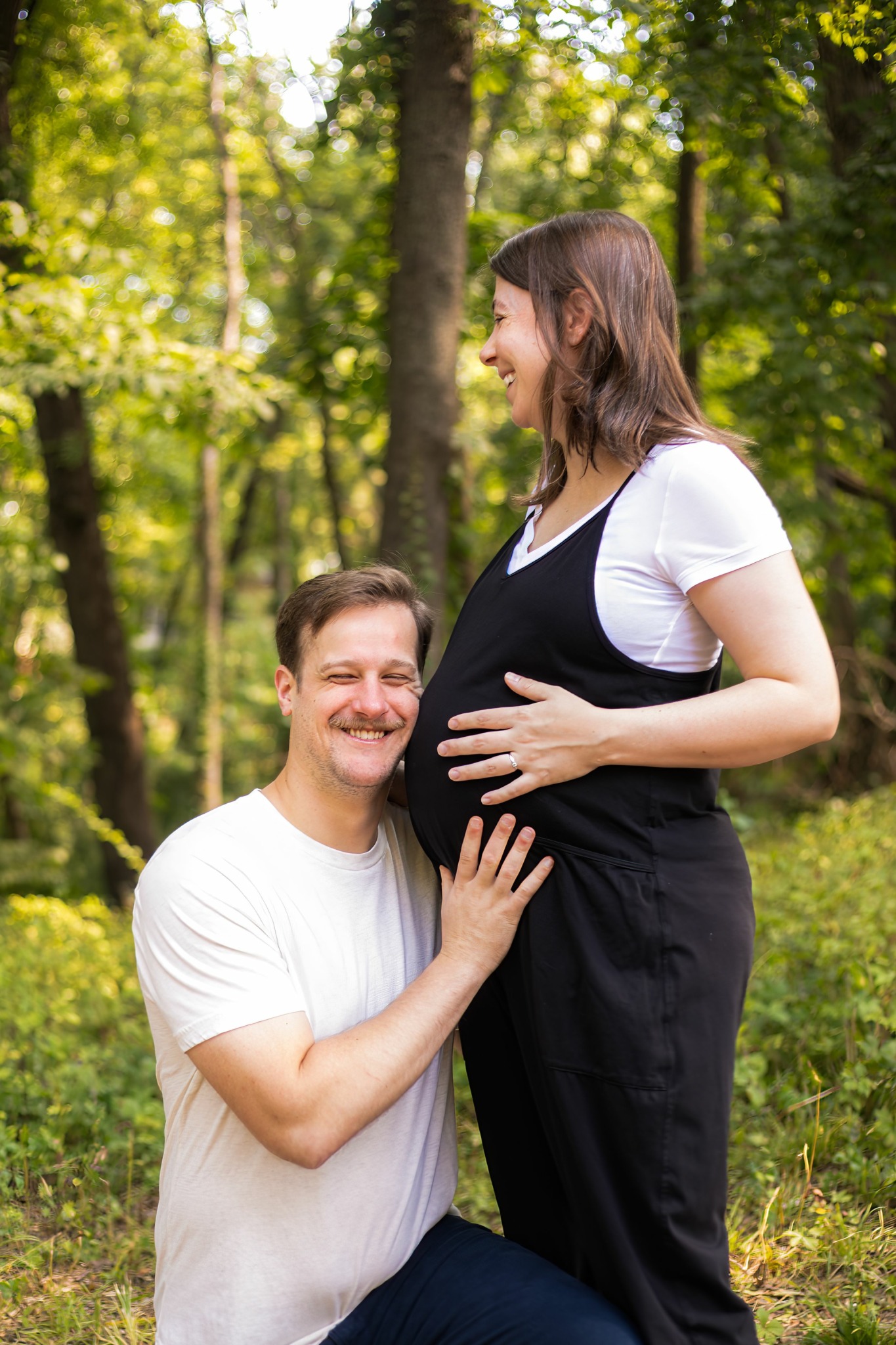 A smiling man and pregnant woman in a forest, with the woman holding her belly, both looking at each other.