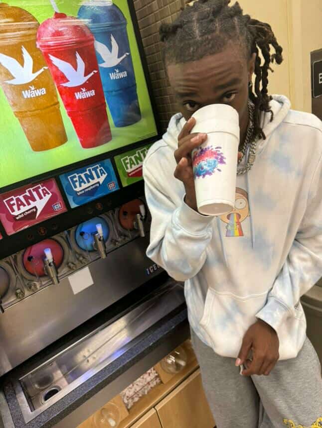 Child with dreadlocks drinking from a cup at a soda fountain, with colorful beverage bottles in the background.