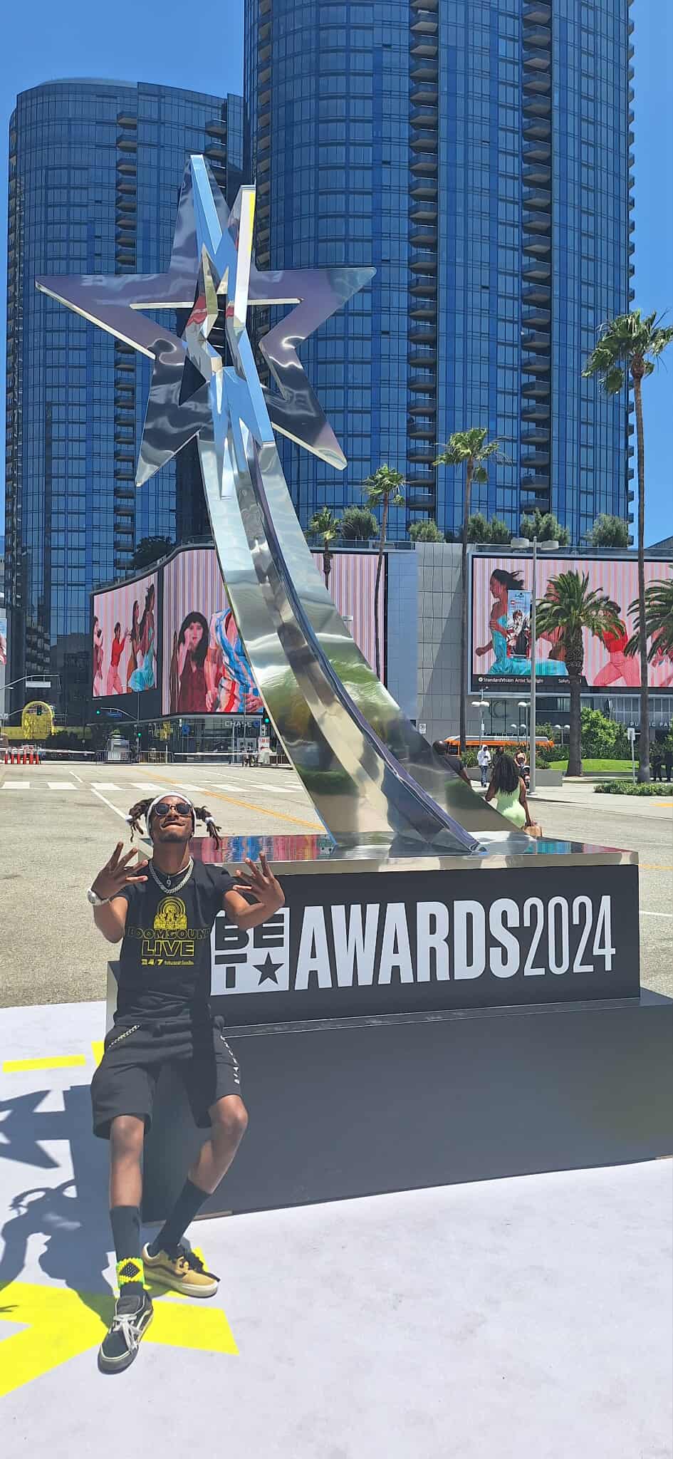 Person standing in front of a large award sculpture with city buildings and palm trees in background.