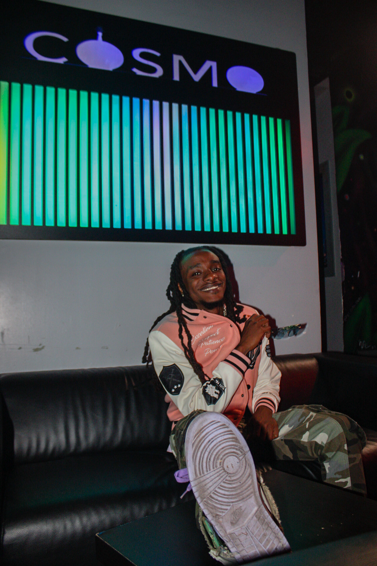 Person with dreadlocks smiling, sitting on a black couch, holding a microphone, in front of a colorful LED display and a sign reading 'COSMO'.