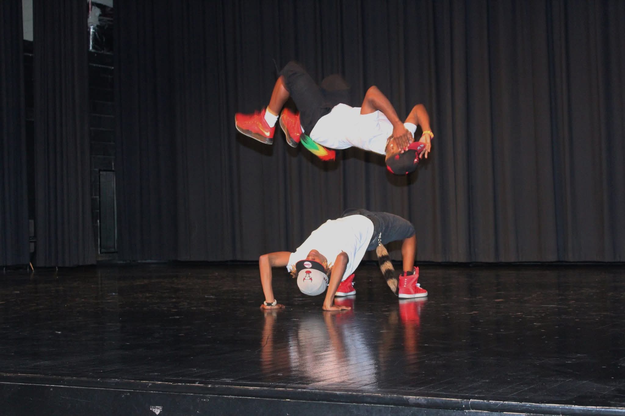 Two people performing acrobatic stunts on stage with a black curtain background, one balancing on hands and the other upside down.