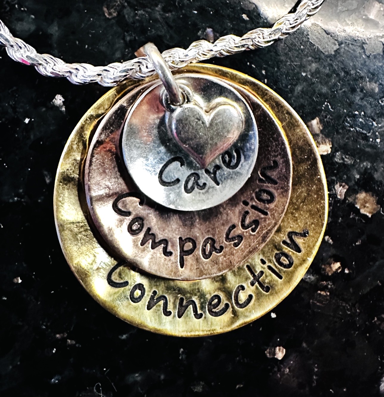 Two layered circular pendants with engraved words, heart symbol, and a rope necklace, on a dark surface.