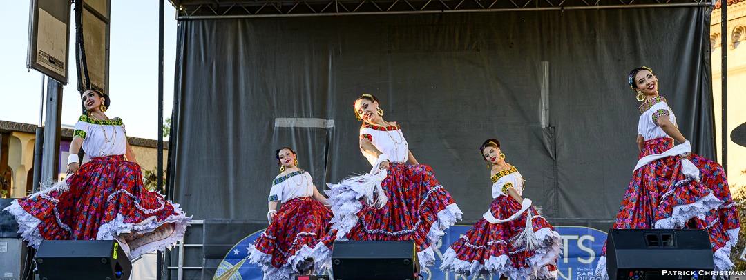 Group of dancers in traditional costumes performing on stage with a black backdrop.