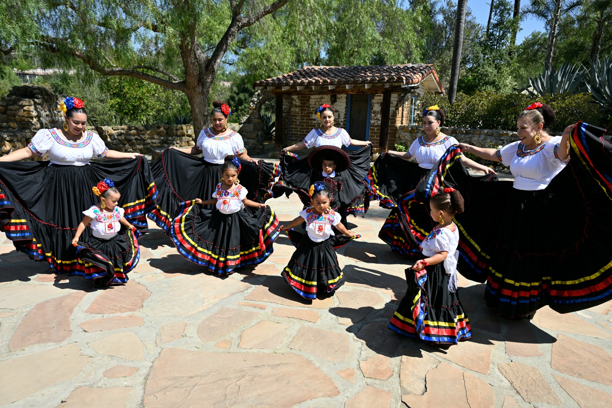 Group of women and children in traditional dresses performing outdoors under trees, with a small wooden structure in background.