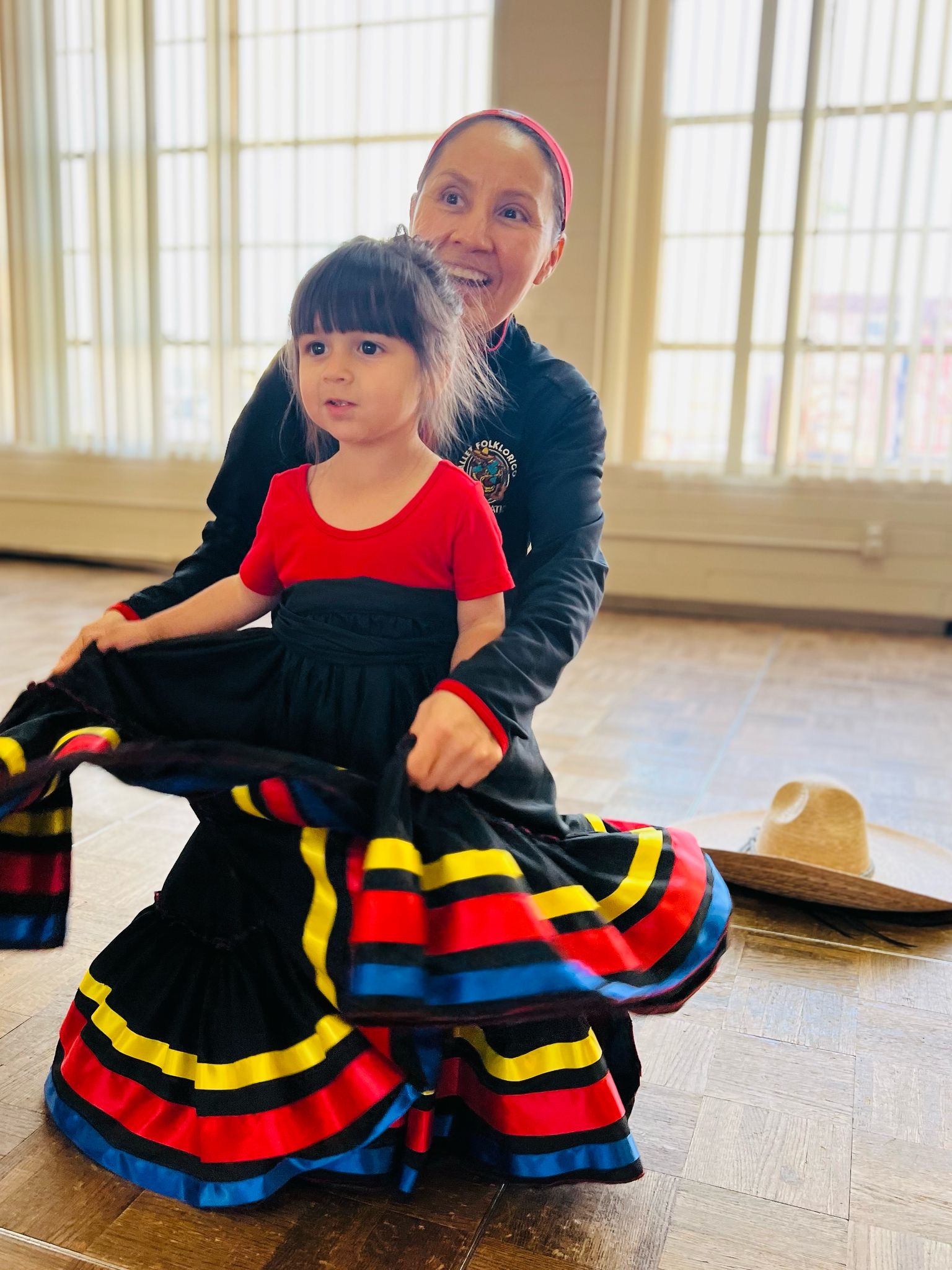Woman and girl in colorful dresses sitting on a large rope, with windows and a hat in the background.