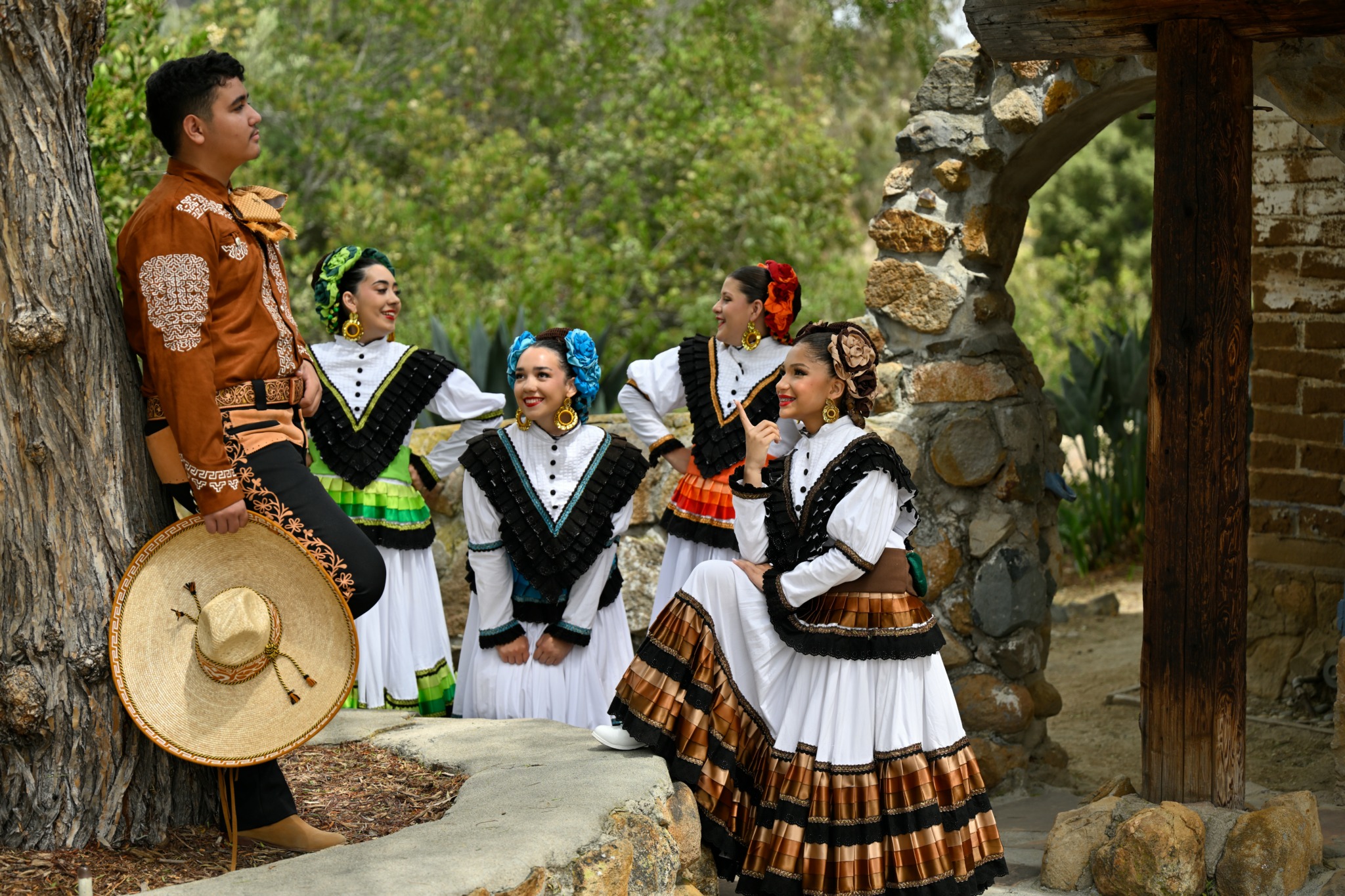 Group of five women in traditional dresses standing outdoors near a stone structure, with a man in traditional attire nearby, trees in background.