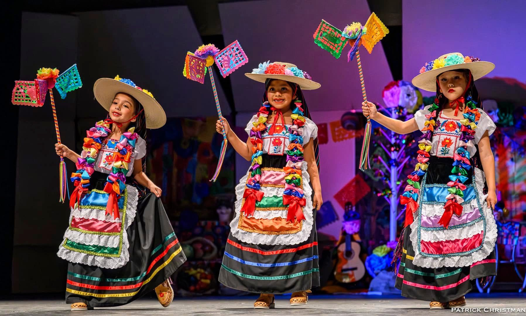 Three women in colorful traditional dresses and wide-brimmed hats perform on stage holding decorated sticks.