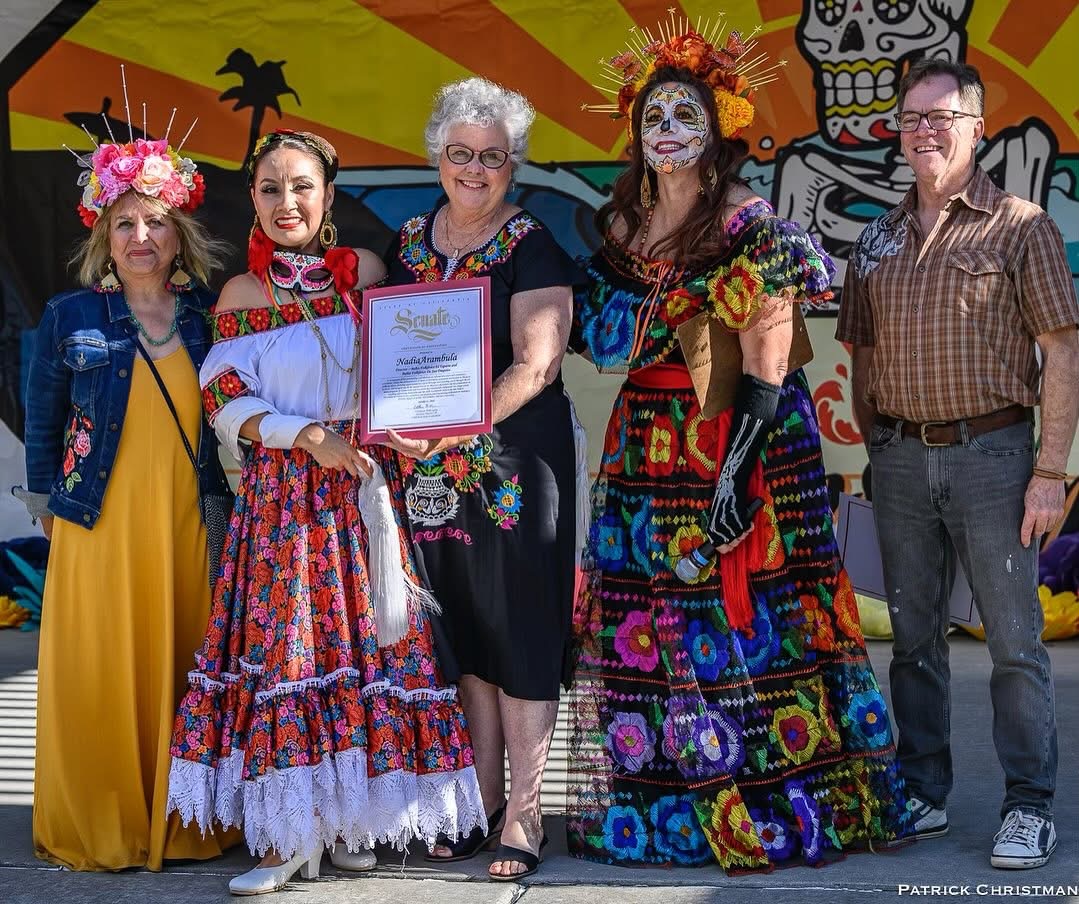 Five people standing together, two women in colorful costumes, one woman holding a framed certificate, background with Mexican-themed decorations.