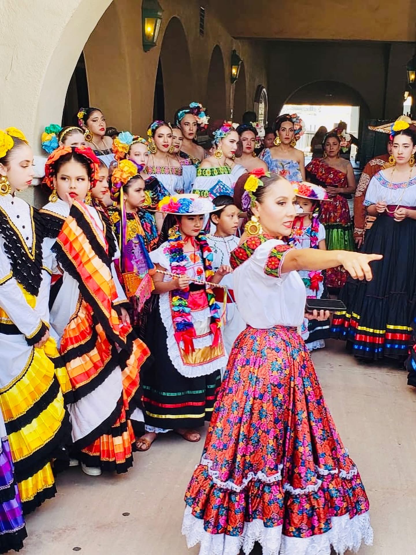 Group of children in colorful traditional dresses, some with flowers in hair, indoors with arched windows and warm lighting.