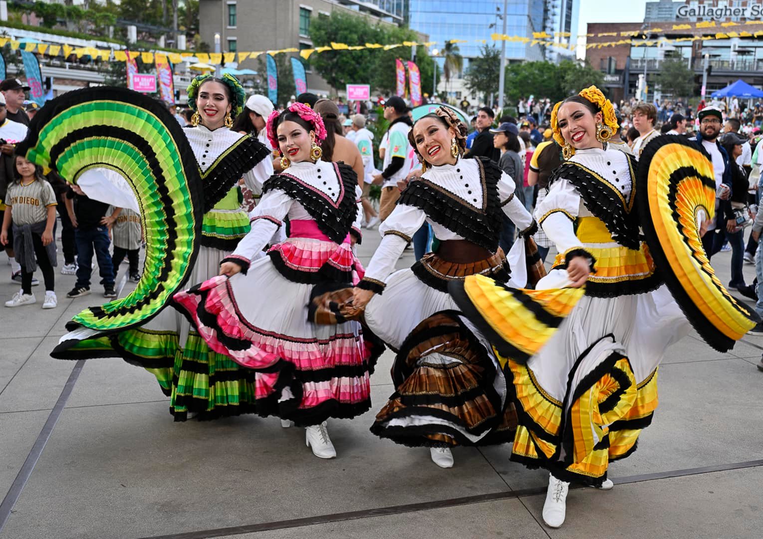 Group of women dancing in colorful traditional dresses at an outdoor event with spectators and decorations.
