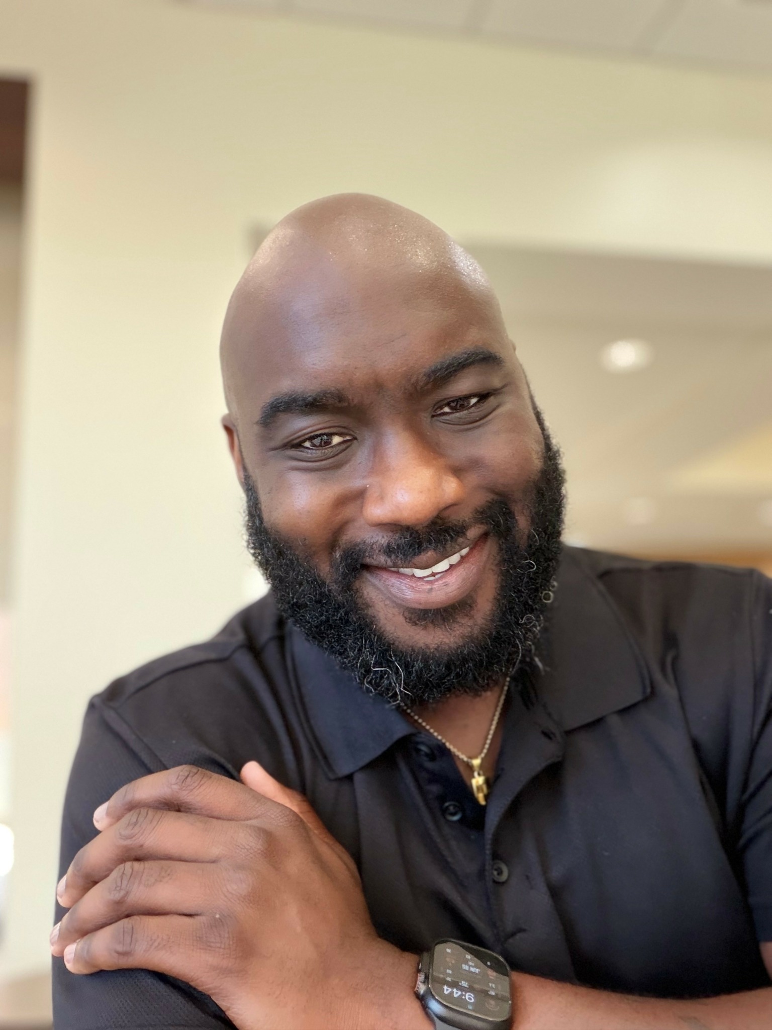 A smiling man with a beard and bald head, wearing a black shirt, crossing his arms.