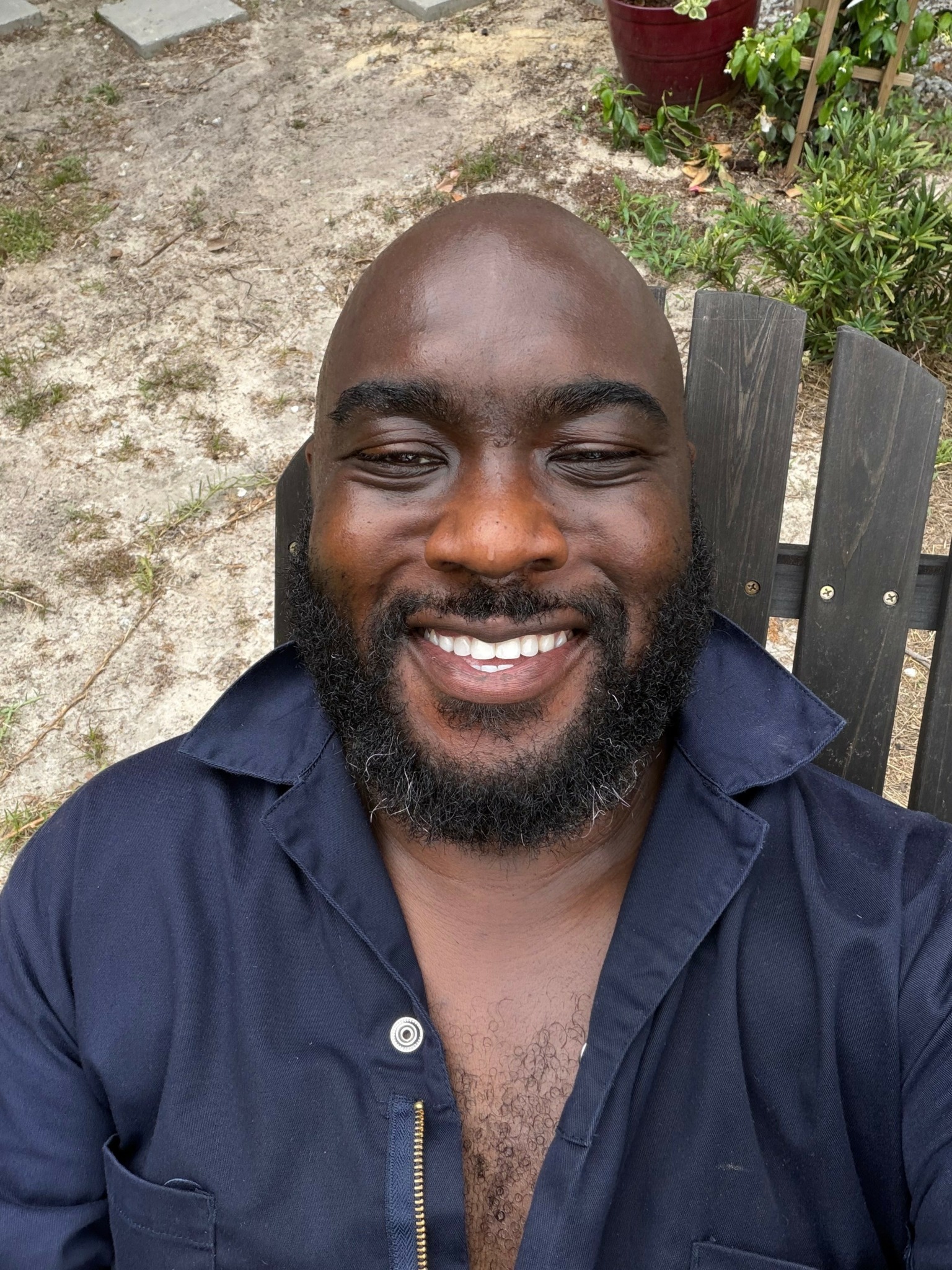 Smiling man with a beard and shaved head outdoors, wearing a dark blue shirt, sitting on a wooden chair.