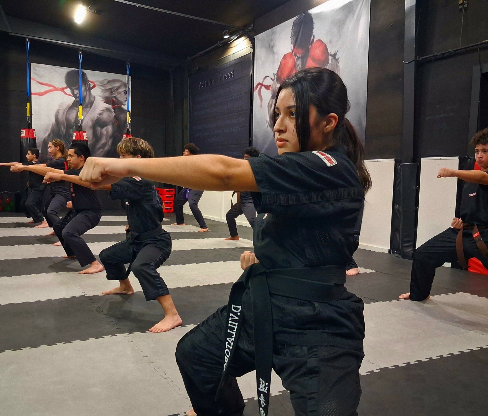 Group of people practicing martial arts in a dojo, focusing on a woman in the foreground with outstretched arm.