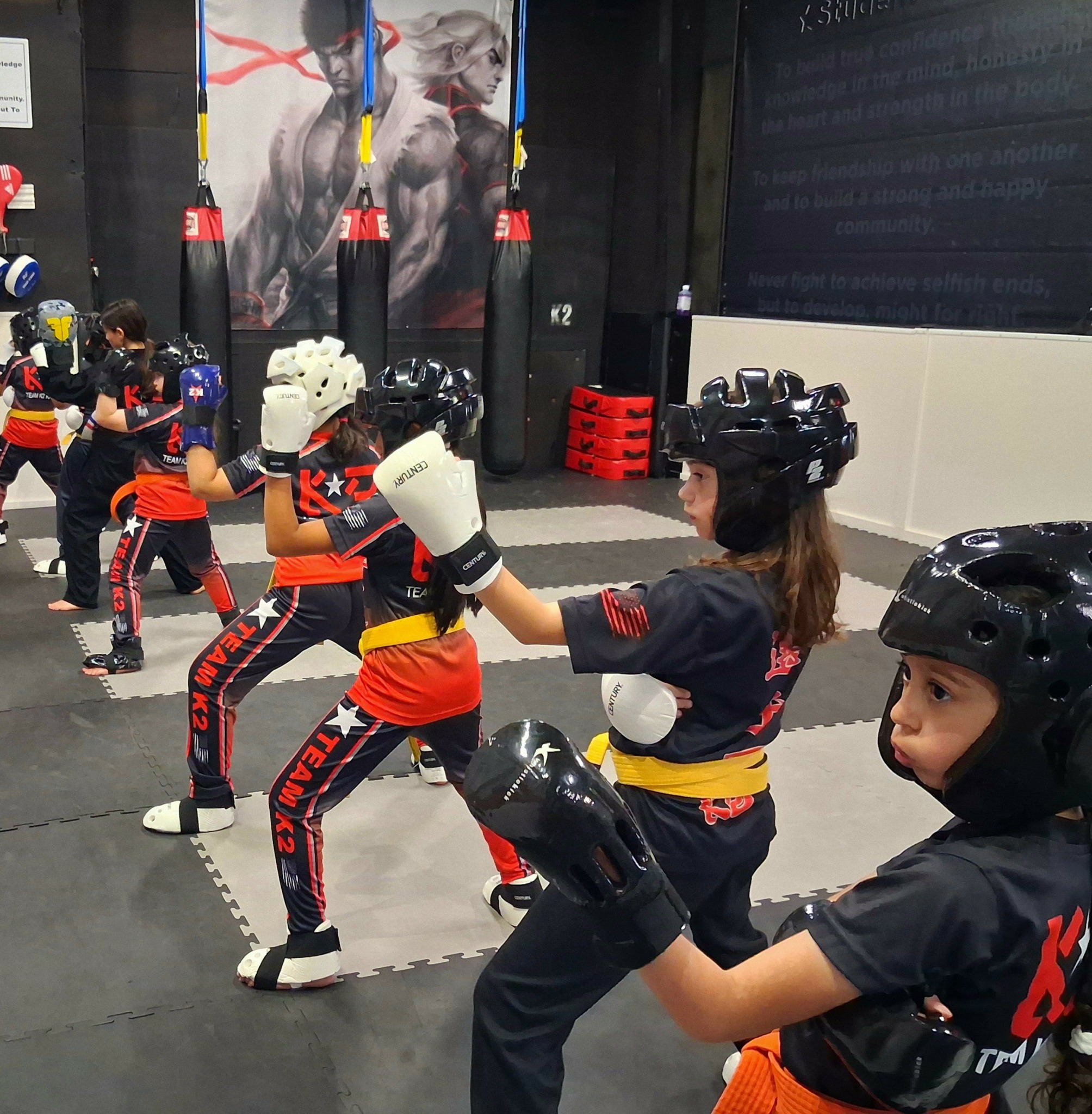 Group of children practicing martial arts in a gym, wearing protective gear and uniforms, with a large poster in the background.