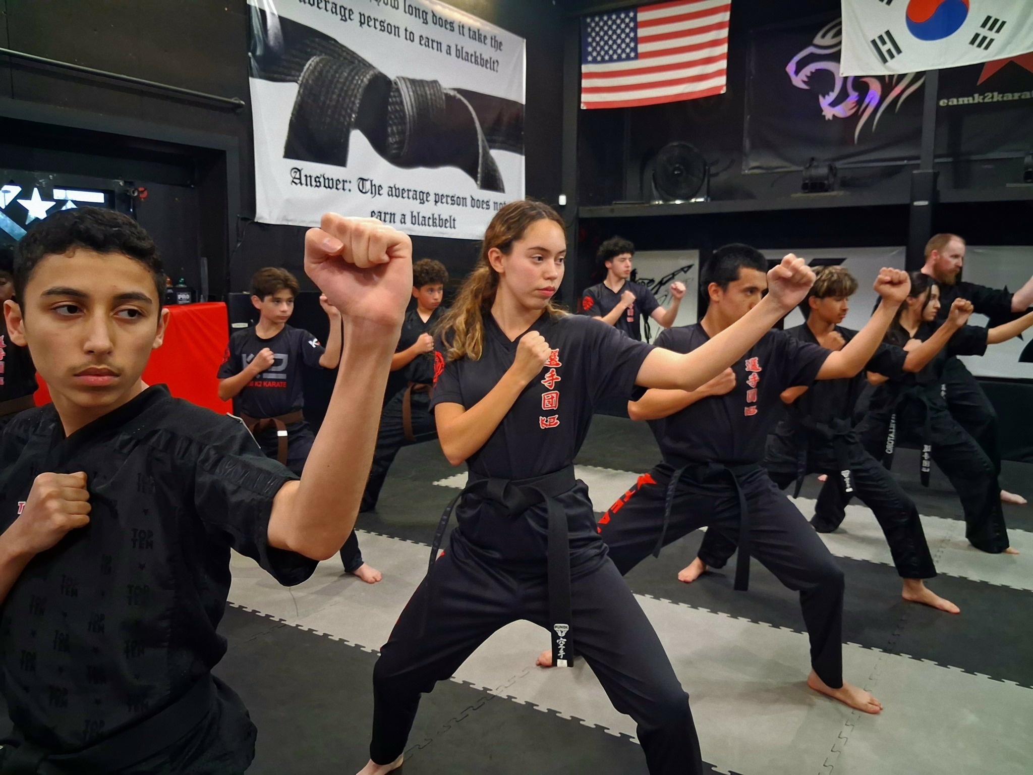 Group of people practicing martial arts in a dojo with flags and banners, some with fists raised, focused on training.