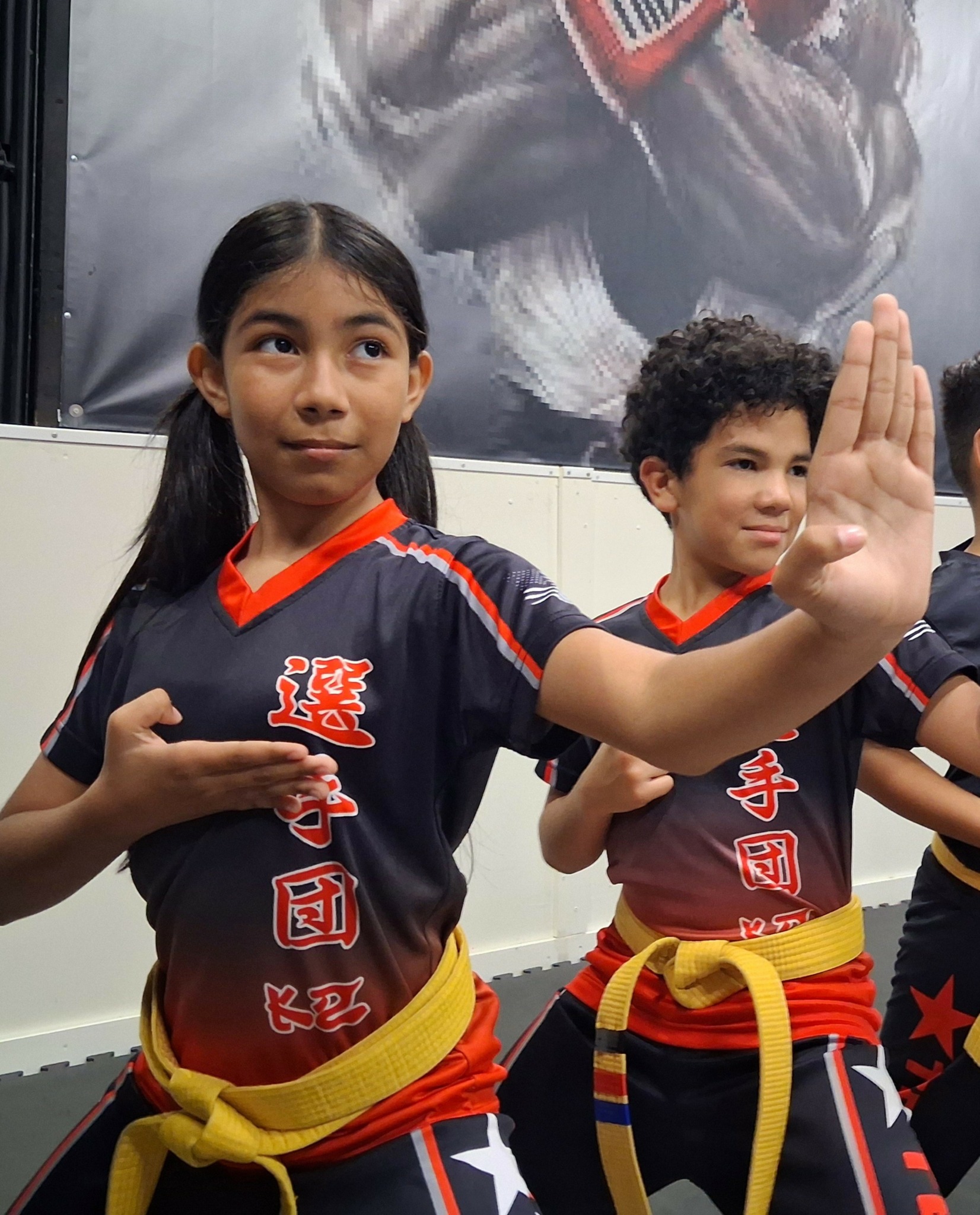 Two children in martial arts uniforms with yellow belts, one girl with long dark hair and one boy with curly hair, standing indoors.