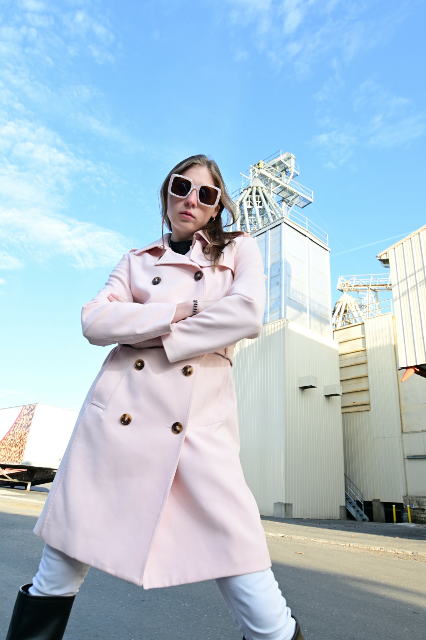 Woman in a pink coat and sunglasses standing outdoors against industrial buildings and blue sky.
