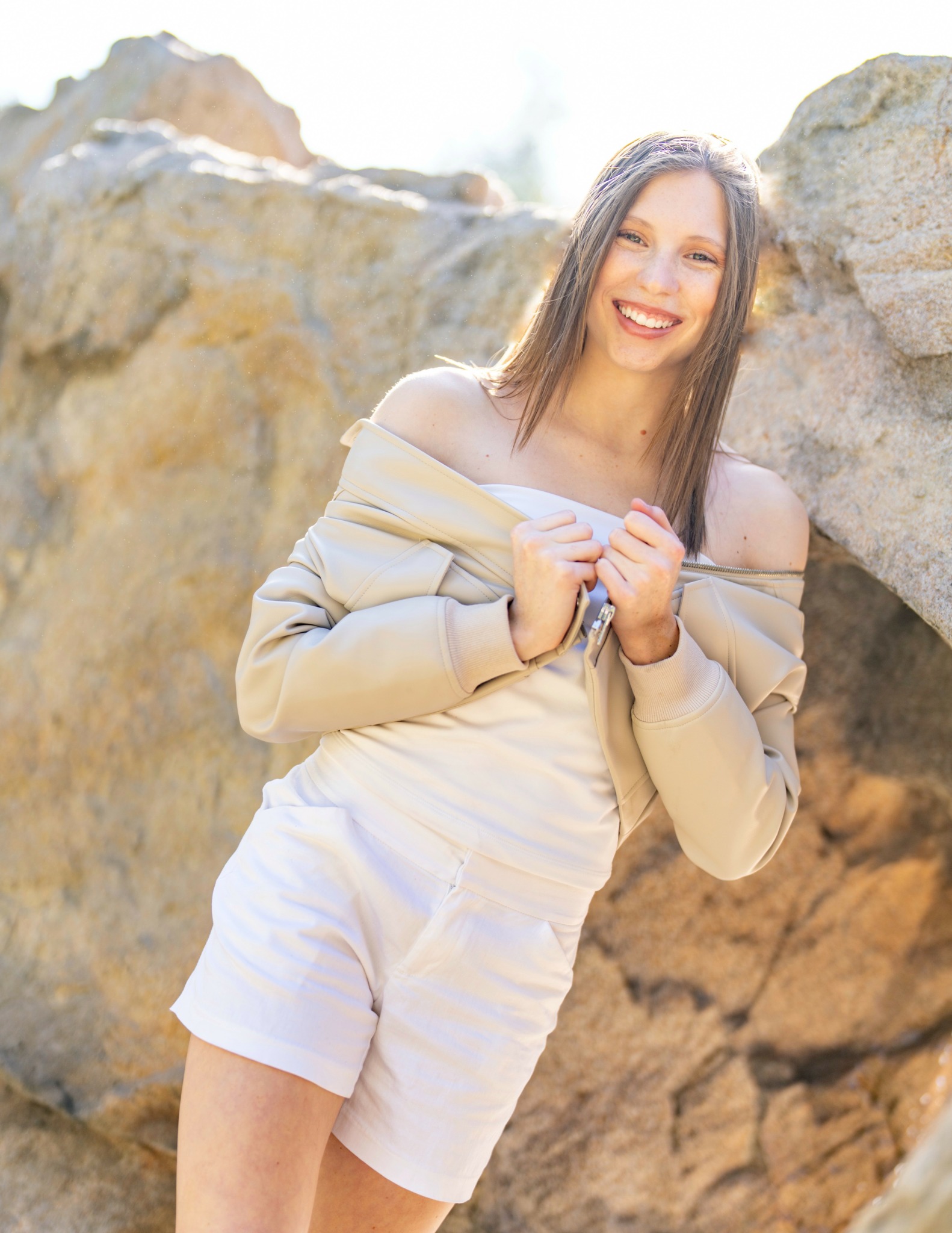 Young woman smiling outdoors, leaning against rocks, wearing a beige off-shoulder top and white shorts.