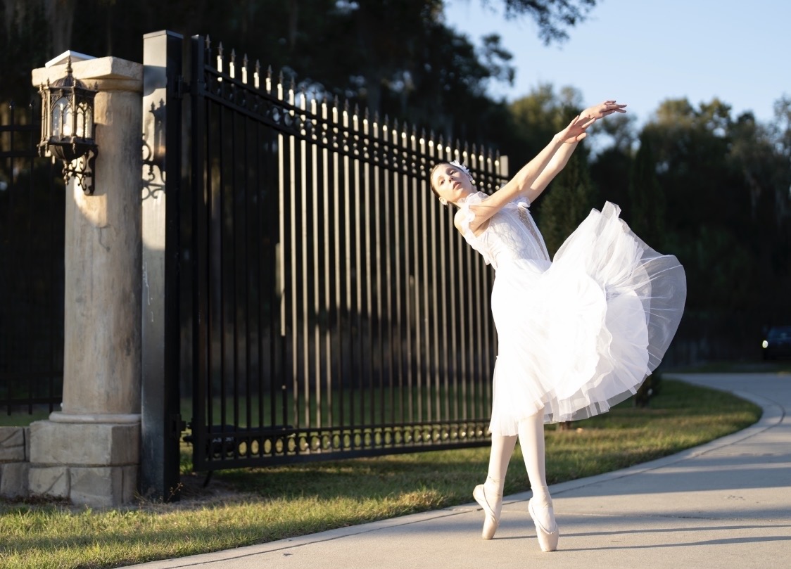 Ballet dancer in white tutu performs outdoors near a black gate, with trees and sky in background.
