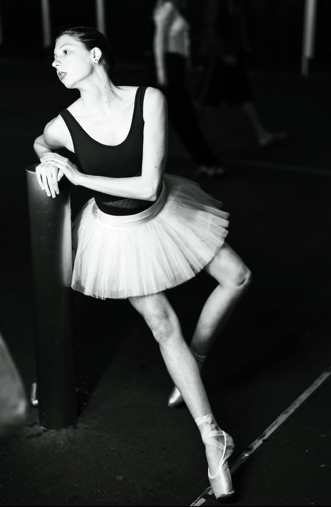 Ballet dancer in tutu and pointe shoes leaning on a pole, in a dance studio, black and white photo.