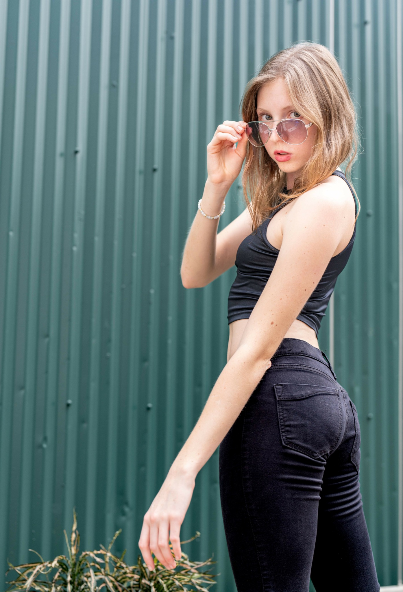 Young woman with sunglasses, wearing a black top and jeans, standing outdoors near a green corrugated metal wall, with one hand adjusting her sunglasses.