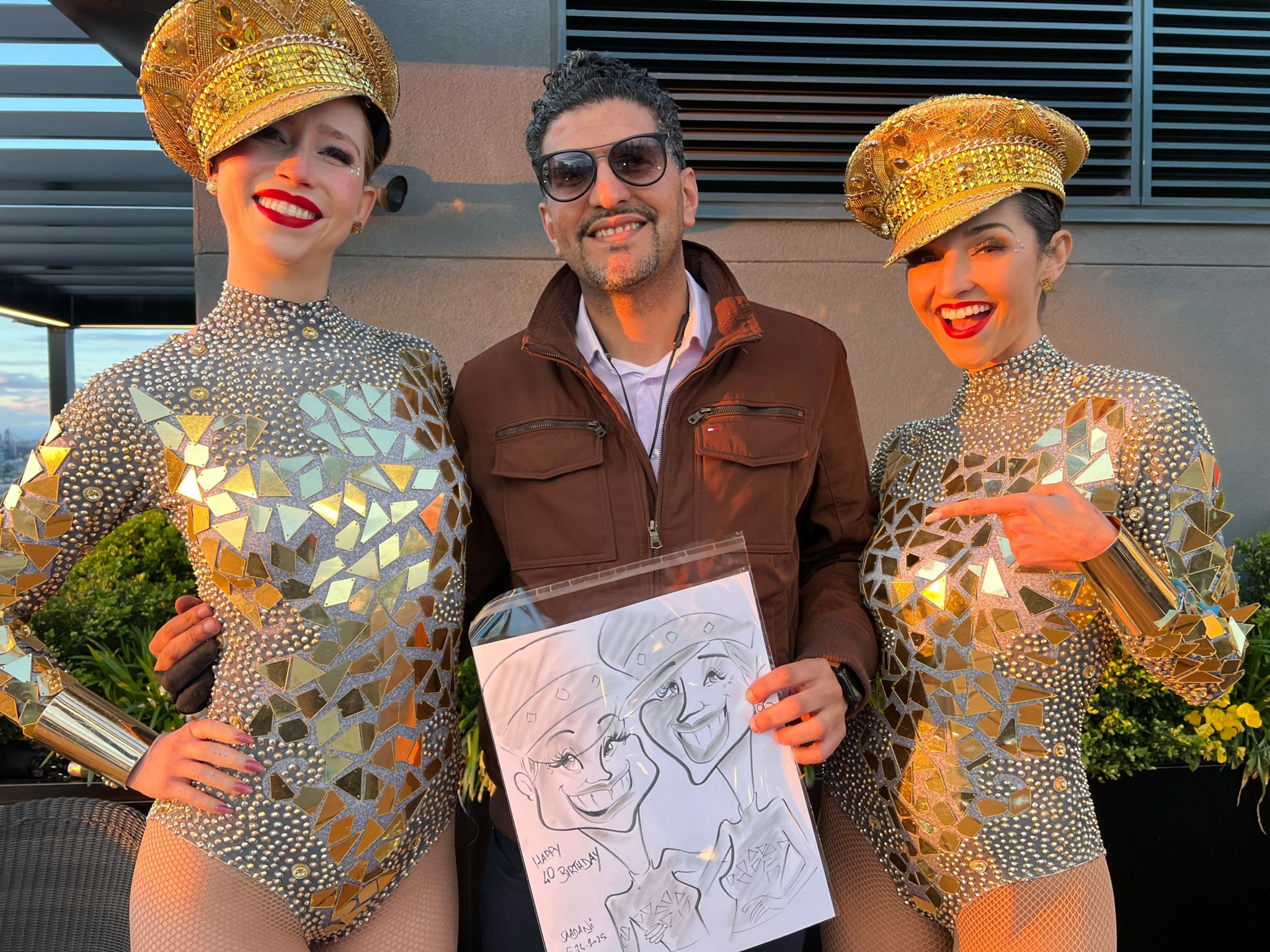 Two women in shiny costumes and a man holding a sketch, all smiling outdoors.