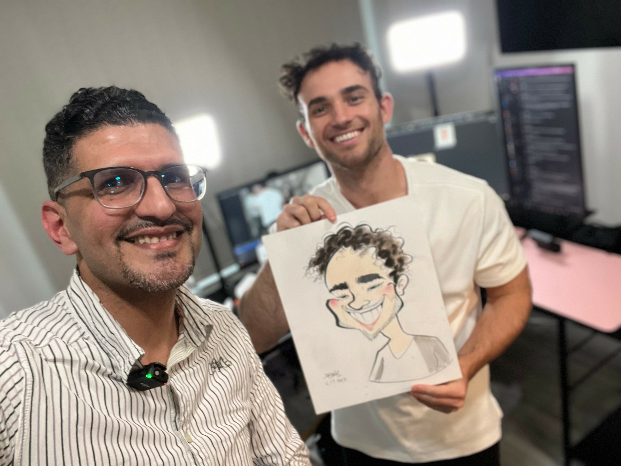Two men smiling, one holding a drawing of a person with curly hair, in a room with computer monitors.