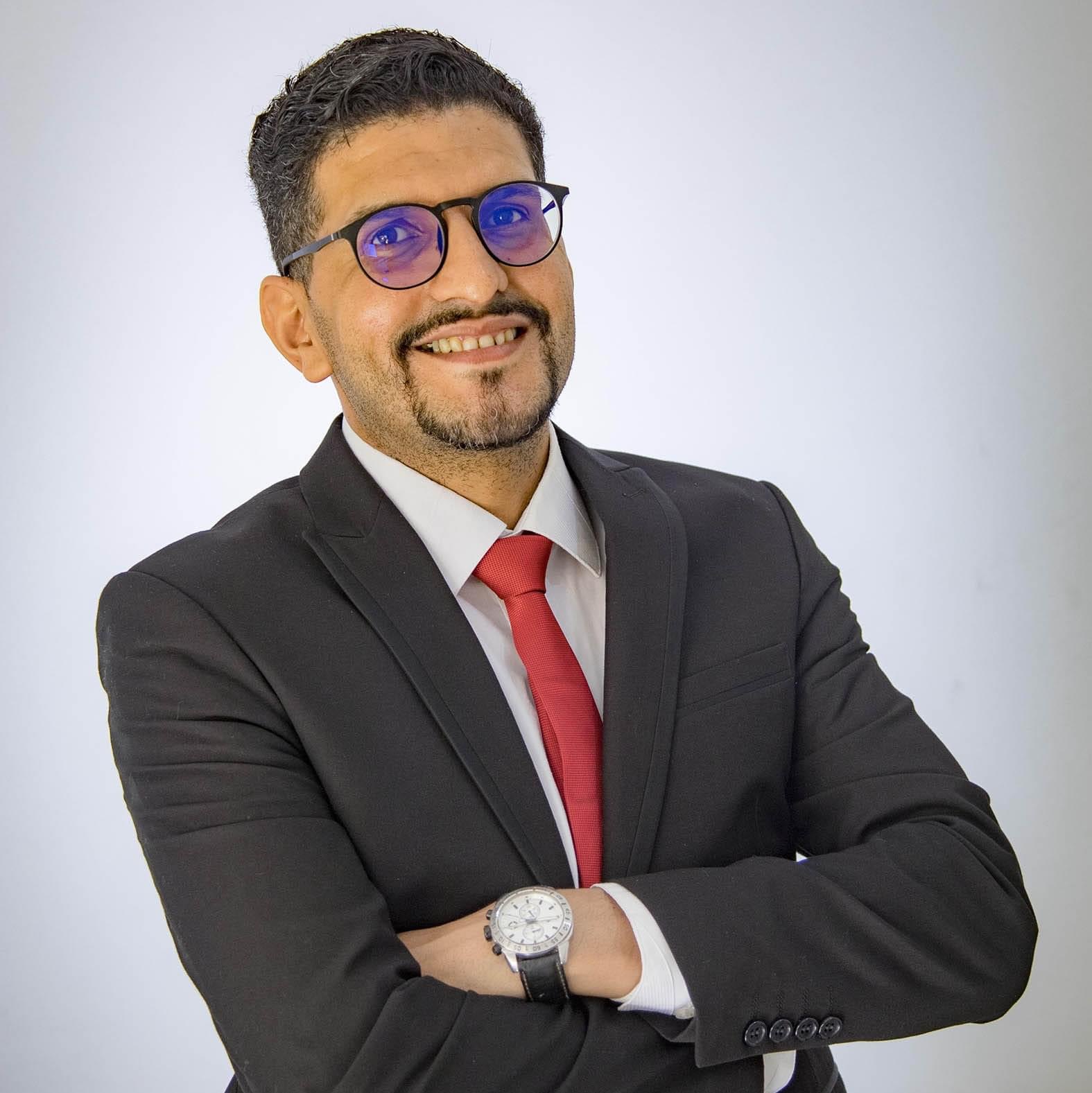 Man in suit with red tie and glasses smiling with arms crossed, white background.
