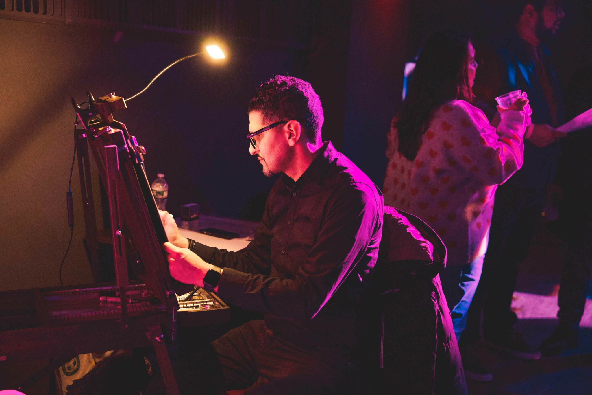 Person playing piano on stage with purple and pink lighting, wearing glasses and dark clothing, in a dimly lit setting.