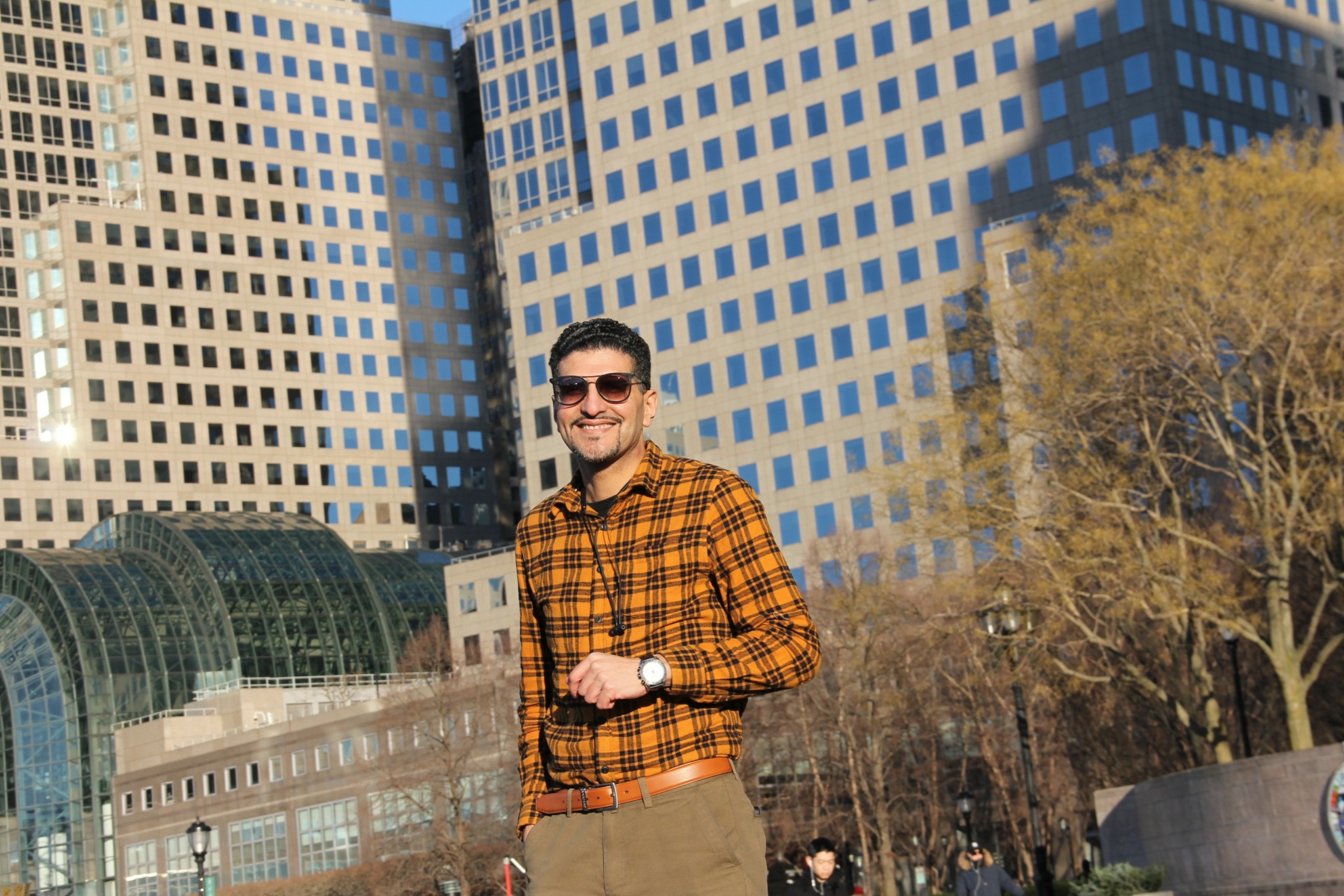 Man wearing sunglasses and a plaid shirt standing outdoors in front of tall buildings and trees.