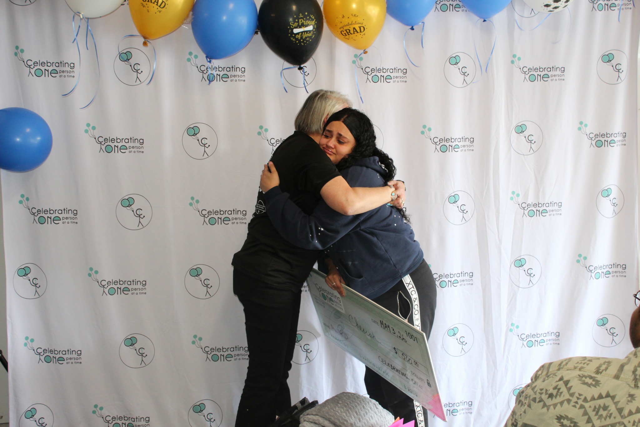 Two people hugging in front of a backdrop with balloons and a banner, one holding a large check or certificate.