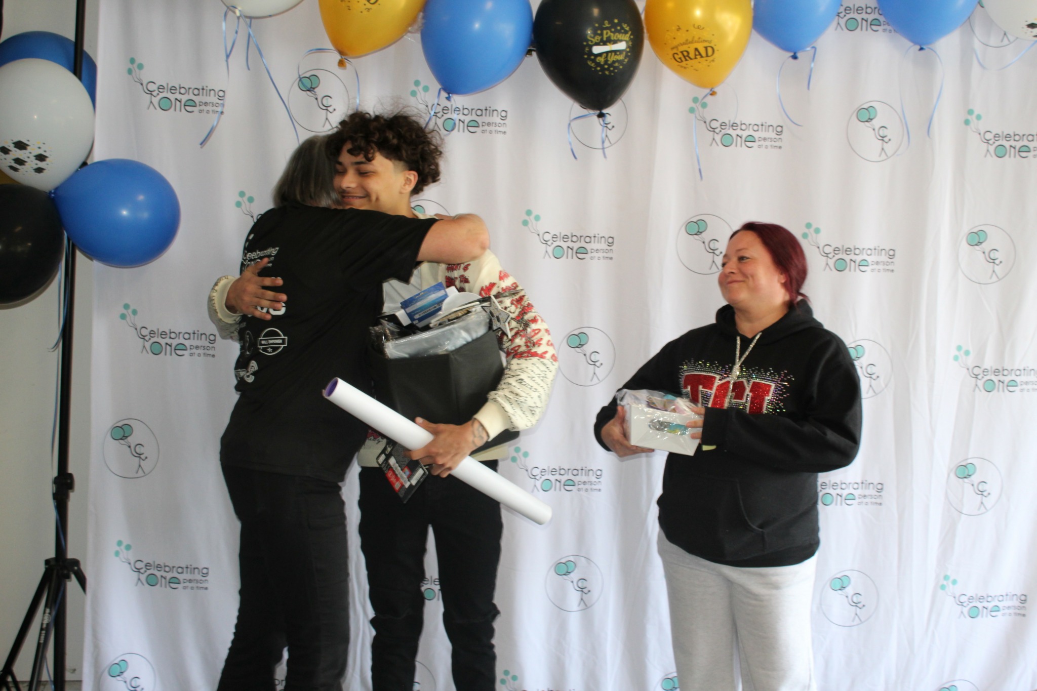 Two women hugging, one holding a rolled paper, surrounded by balloons and a backdrop with logos.