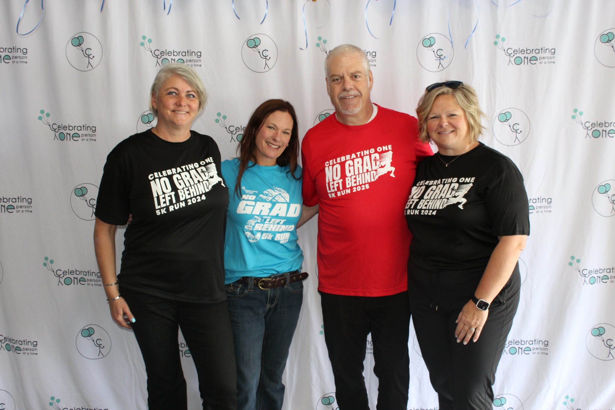 Four people standing together in front of a backdrop, smiling, with two women wearing black T-shirts and two others in blue and red shirts.