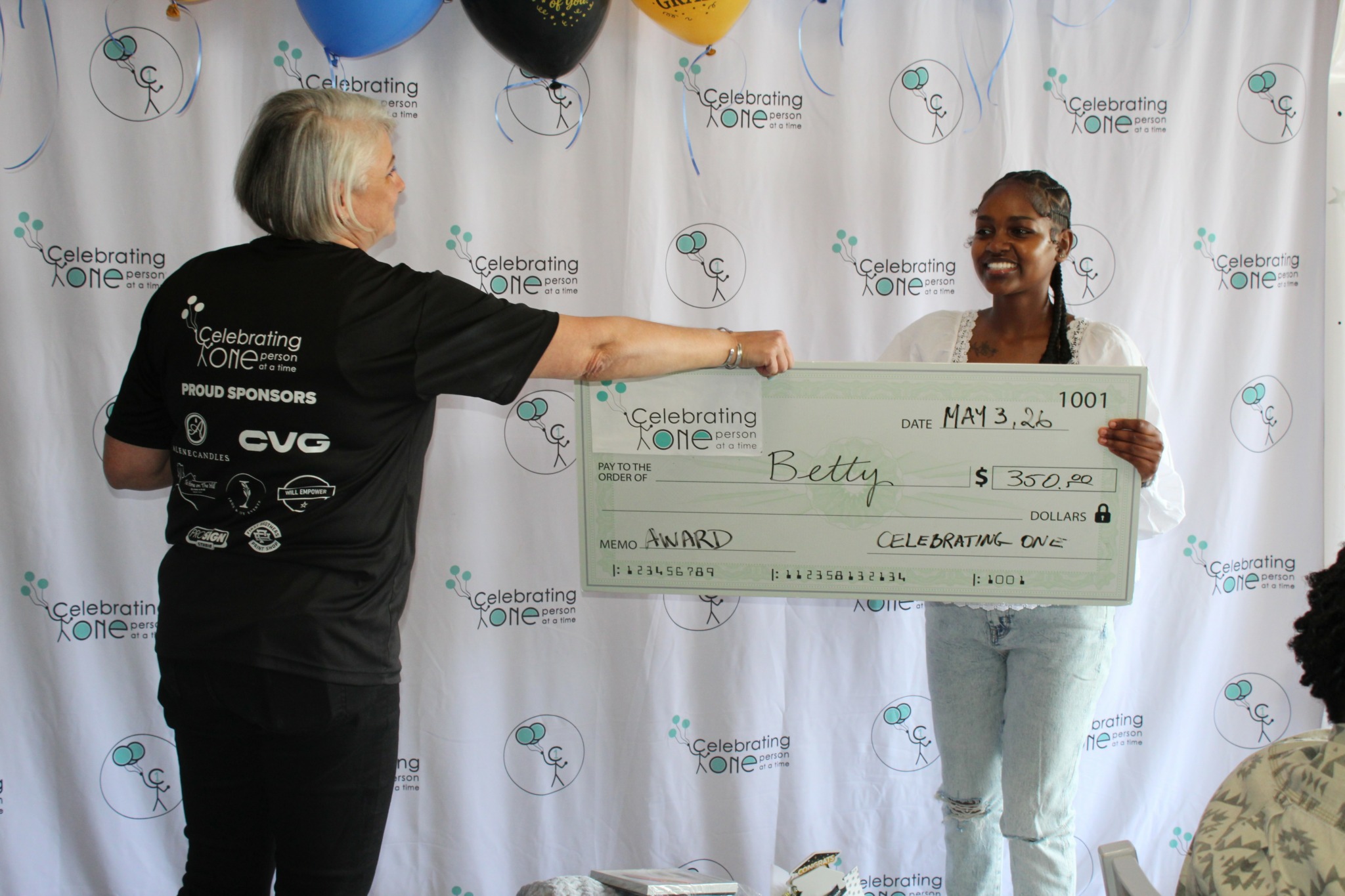 Two women exchanging a large check at an event with a backdrop displaying logos and text, smiling and facing each other.