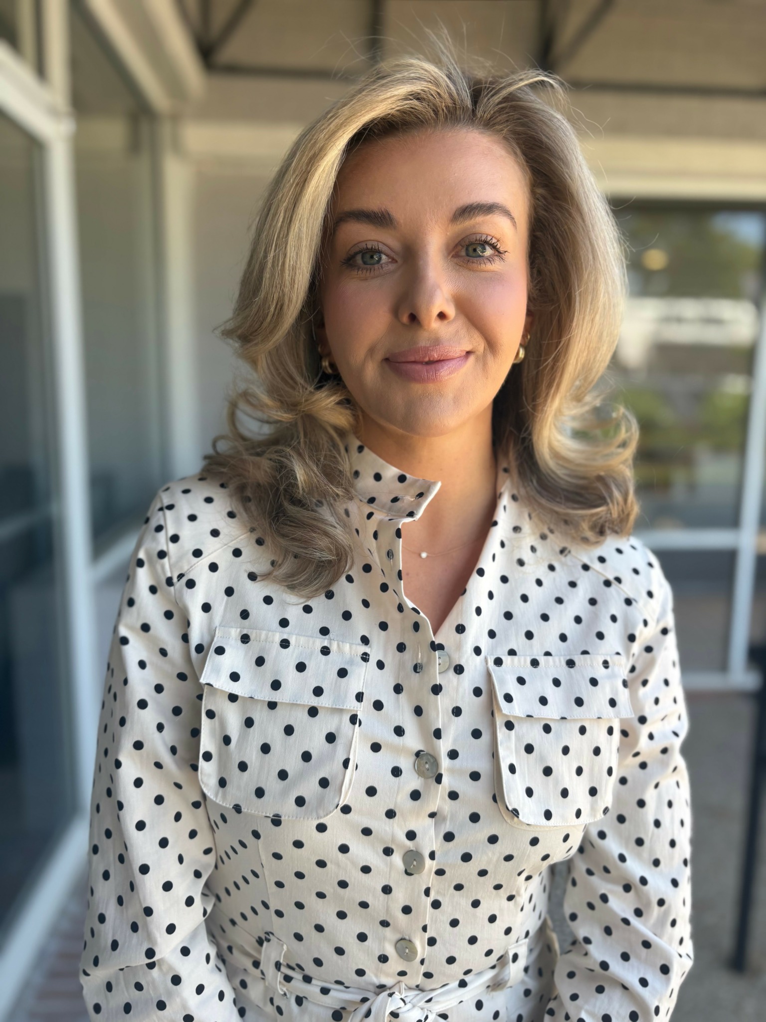 Young woman with wavy blonde hair wearing a white polka dot shirt, standing outdoors near a building with glass windows.