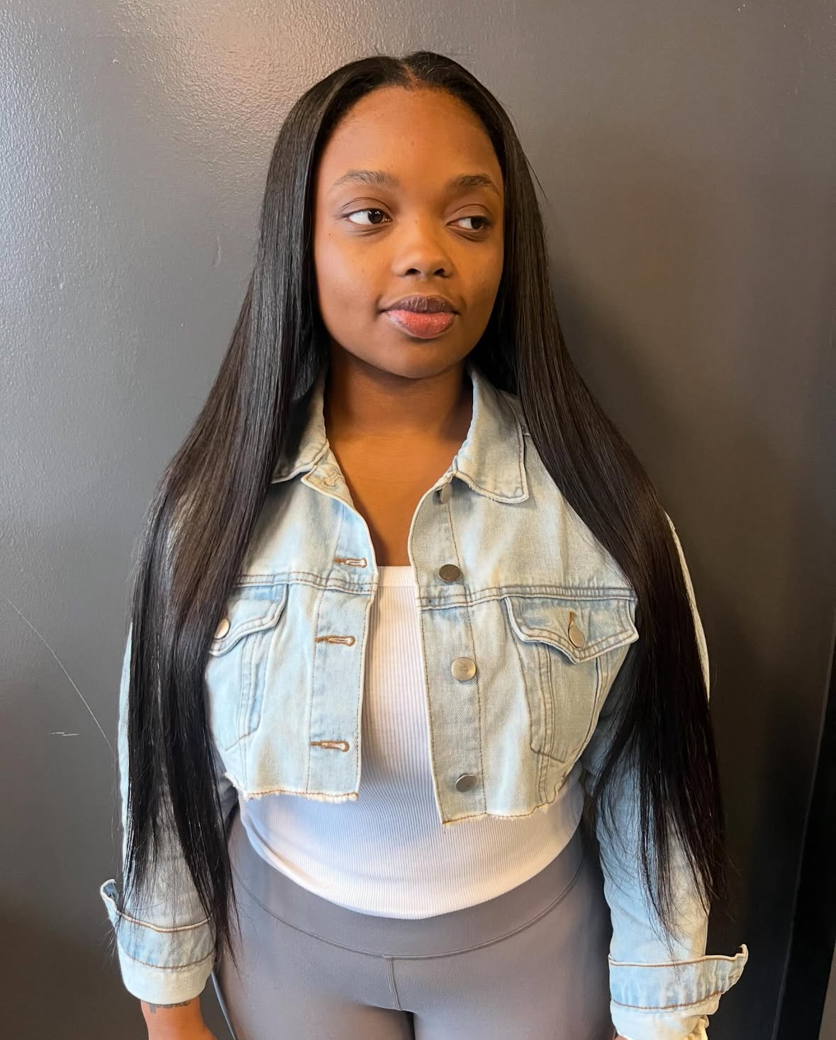 Young woman with long, straight black hair wearing a denim jacket and white top, standing against a gray wall.