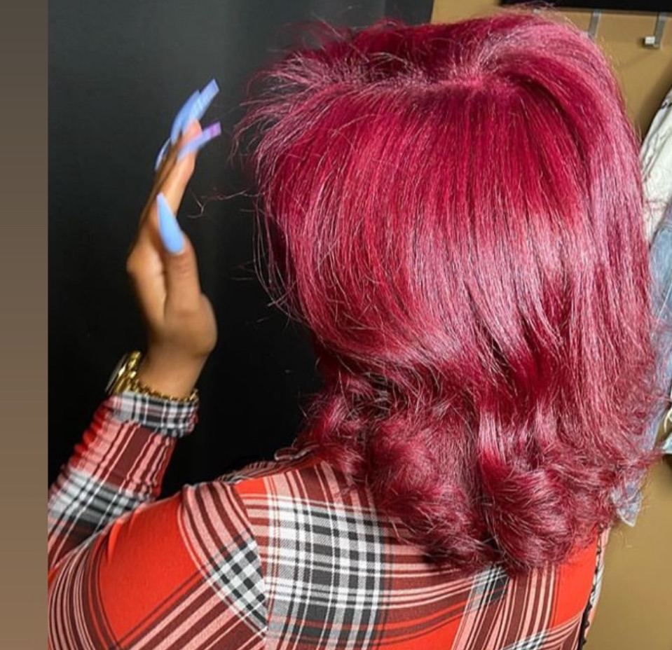 Back view of a person with vibrant pink hair, wearing a plaid shirt, with hand raised showing long blue nails.