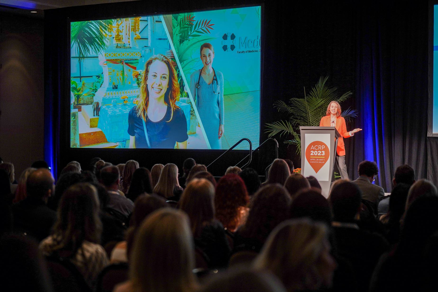 A woman speaks at a podium during a conference, with a large screen behind her displaying colorful images of two women and a pool scene.